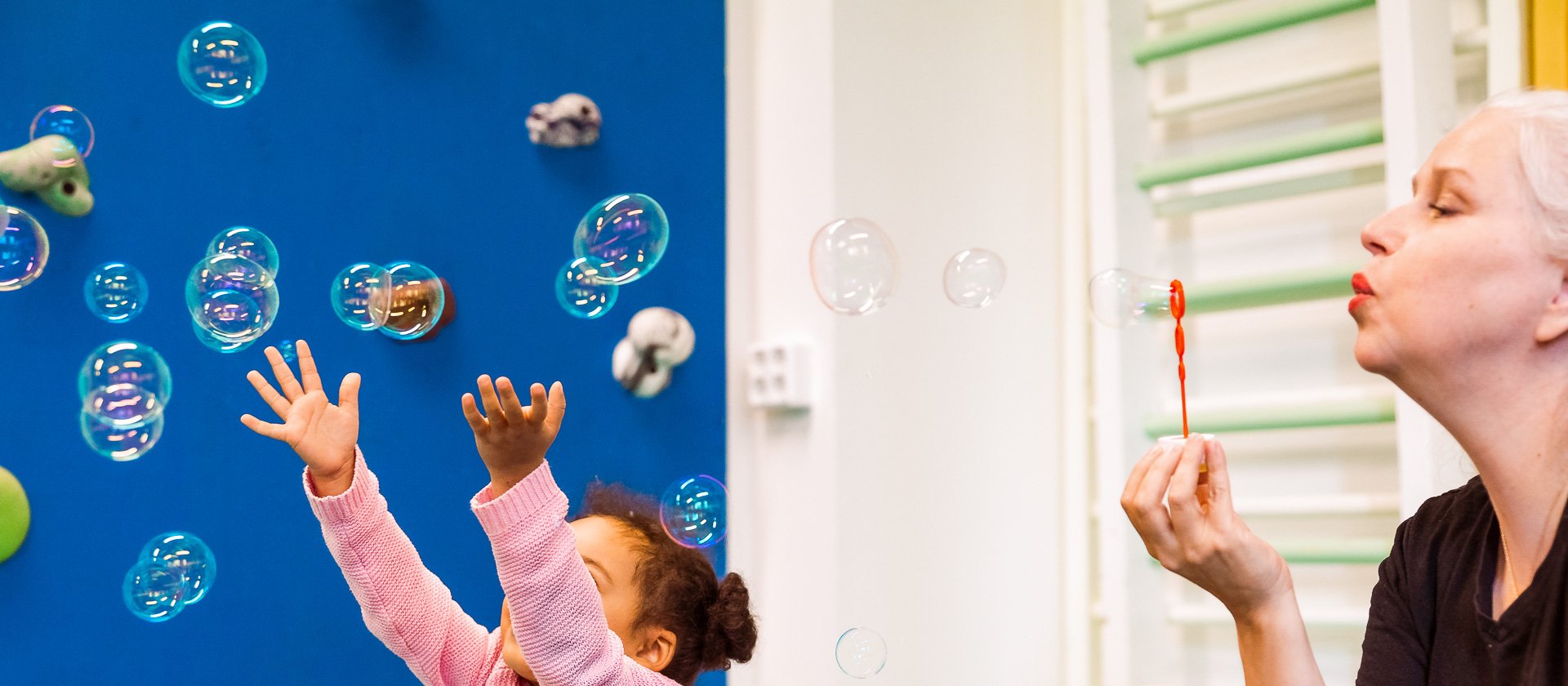 An adult blows bubbles while a child reaches towards them.